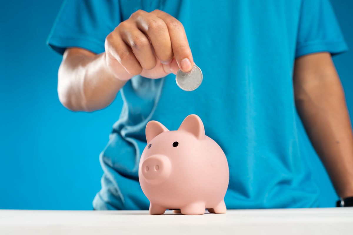 A close-up of an adult's hand dropping a coin into a piggy bank, symbolizing savings and investment.