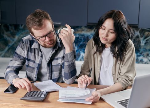 A young couple sitting at a wooden kitchen table reviewing financial documents with a calculator and coffee.
