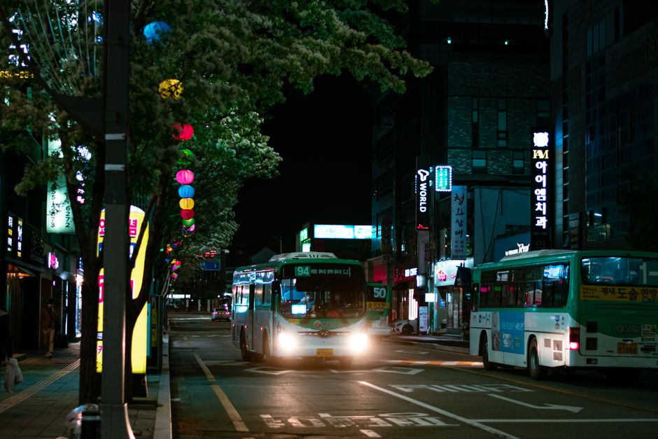 Urban street at night with illuminated buses and colorful neon lights.